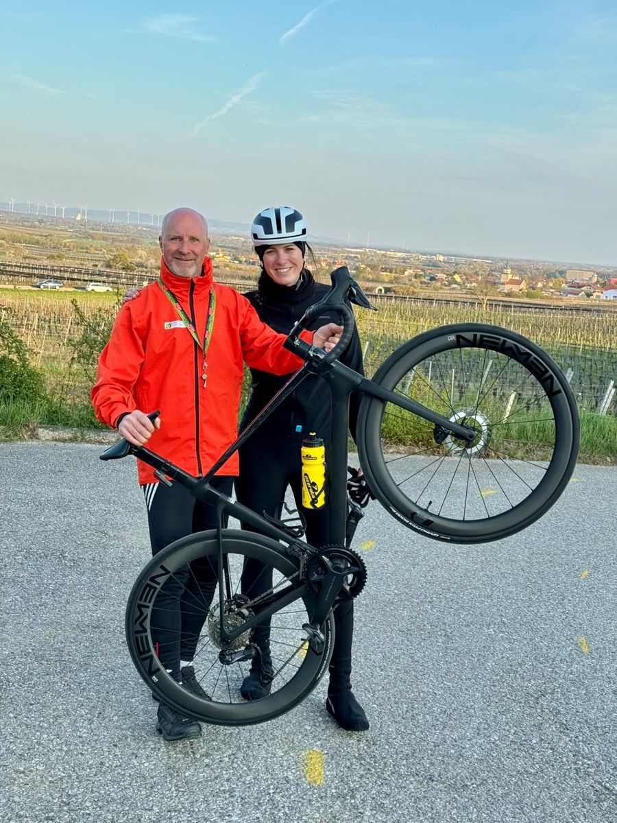 Dani und Roland stehen lachend vor den Weinbergen, Roland hält ein Rennrad in die Höhe