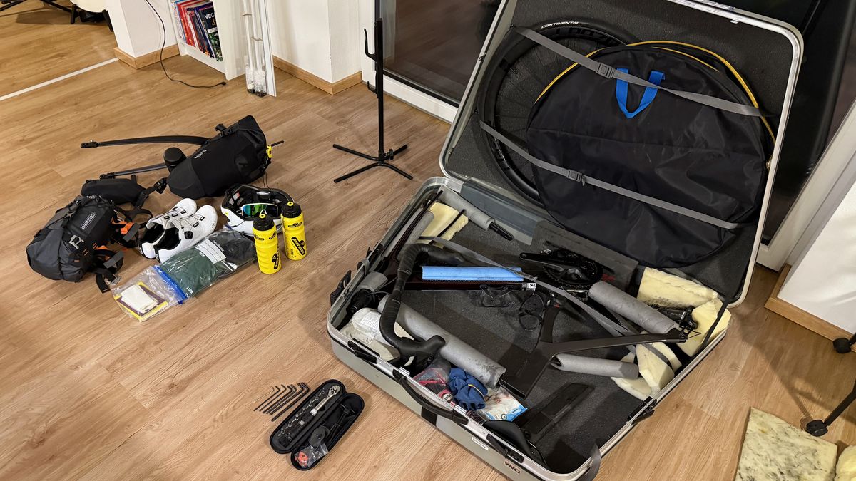 Offener Radkoffer mit dem zerlegten Rad, daneben auf dem Holzboden Taschen, Radschuhe, Flaschen und Werkzeug