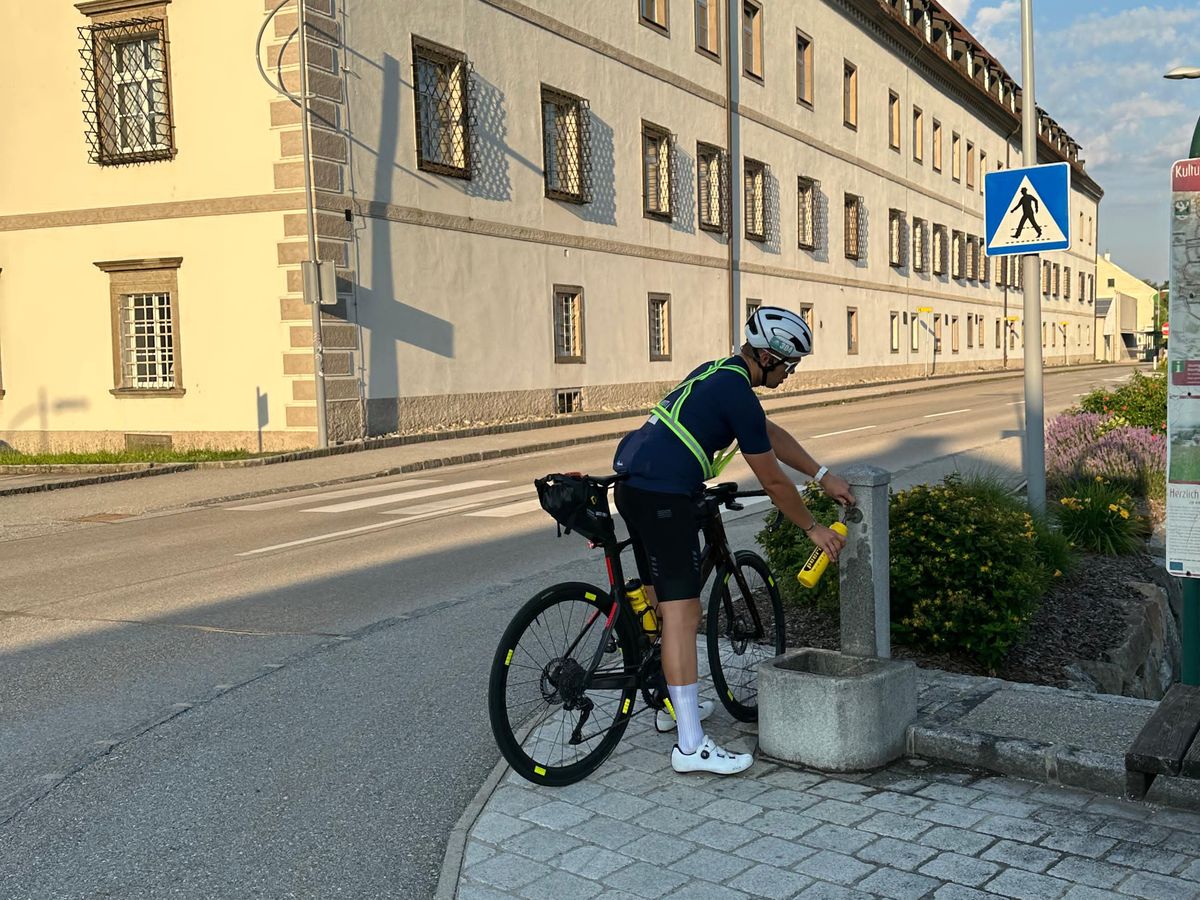 Martin steht mit dem Rad an einem Brunnen vor einem langen gelben Gebäude und füllt eine gelbe Trinkflasche
