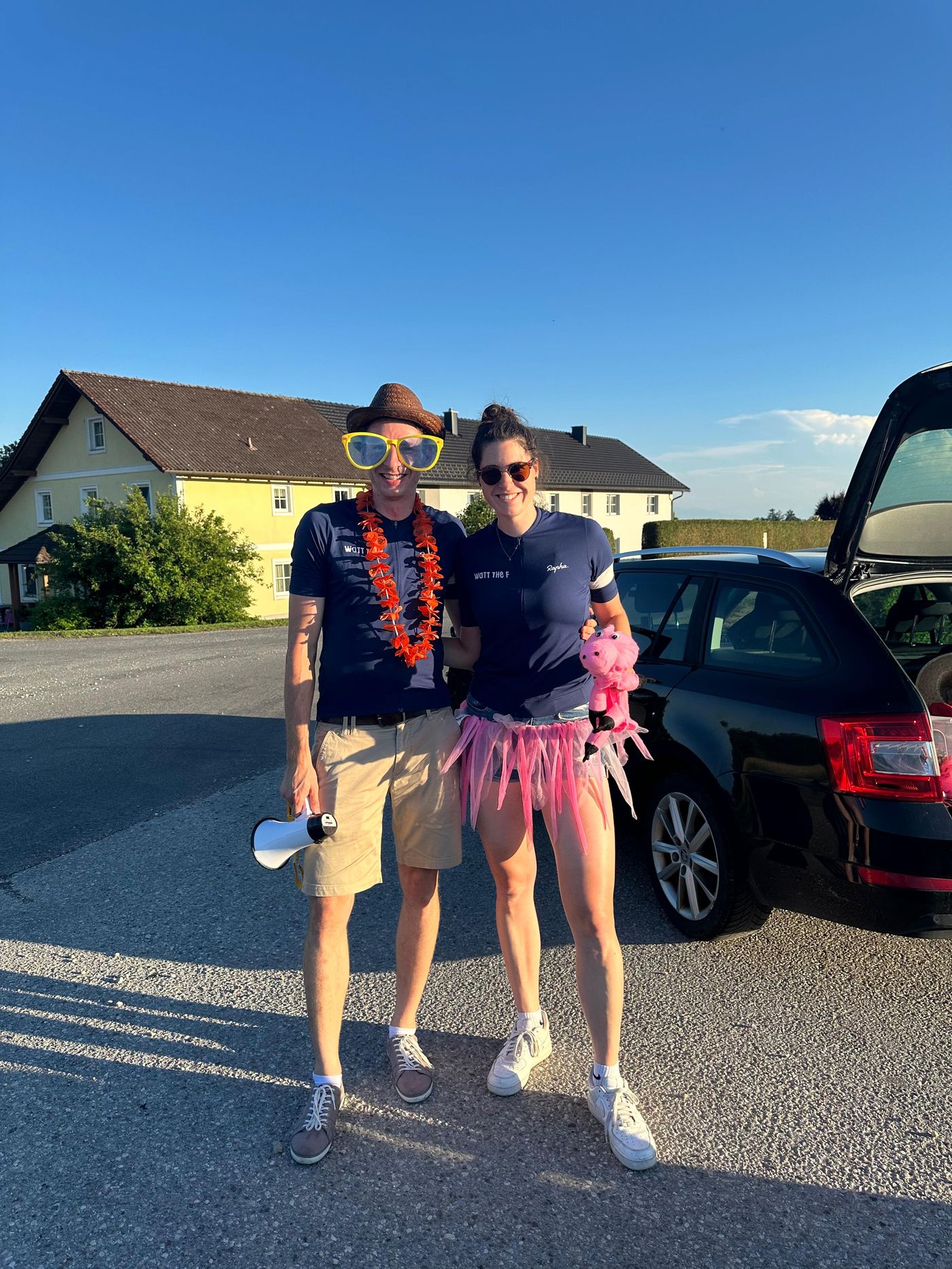 Dani und Andreas in Watt-the-F-Trikots am Straßenrand, Dani im pinken Tüllrock, Andreas mit Cowboyhut, Blumenkette, riesiger Sonnenbrille und Megafon