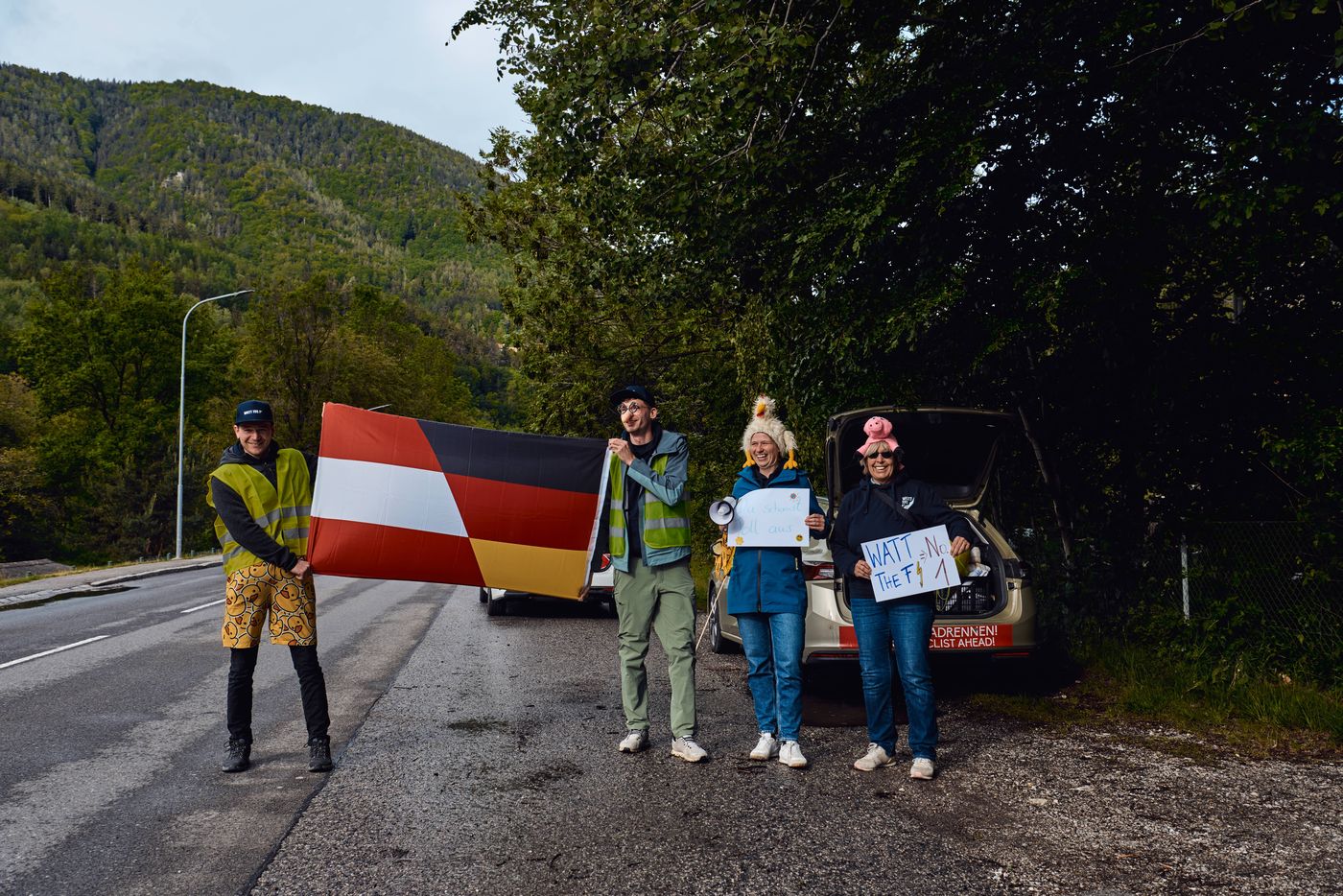 Vier Menschen am Straßenrand mit einer kombinierten Österreich-Deutschland-Fahne und einem Schild