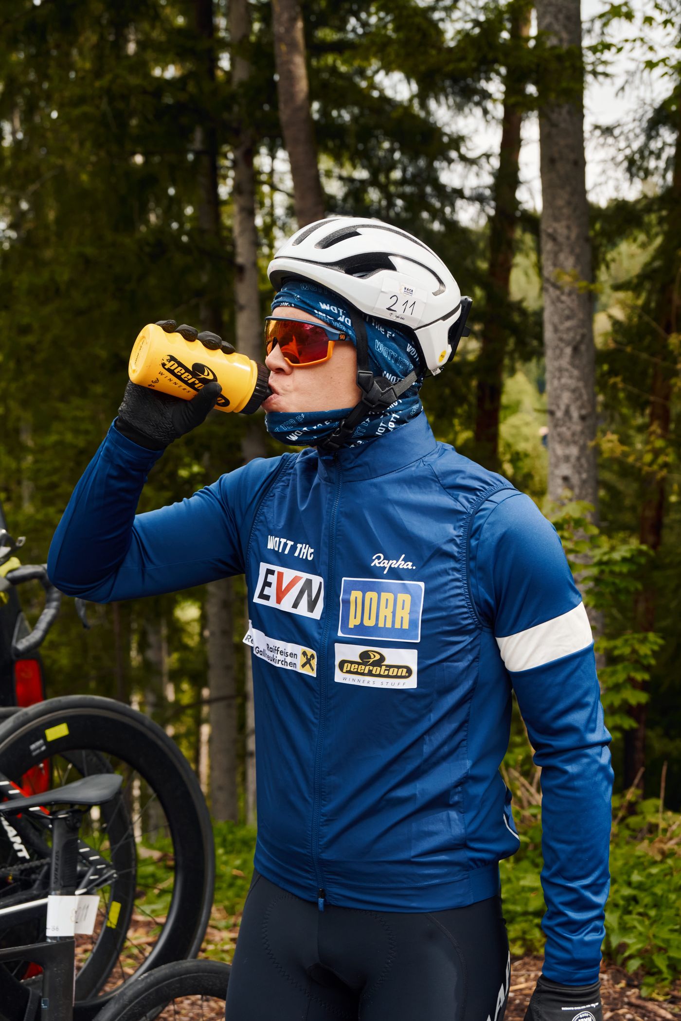 Dani in blauer Jacke mit Helm und Startnummer 211 trinkt aus einer gelben Flasche, neben ihr das Rad am Pacecar