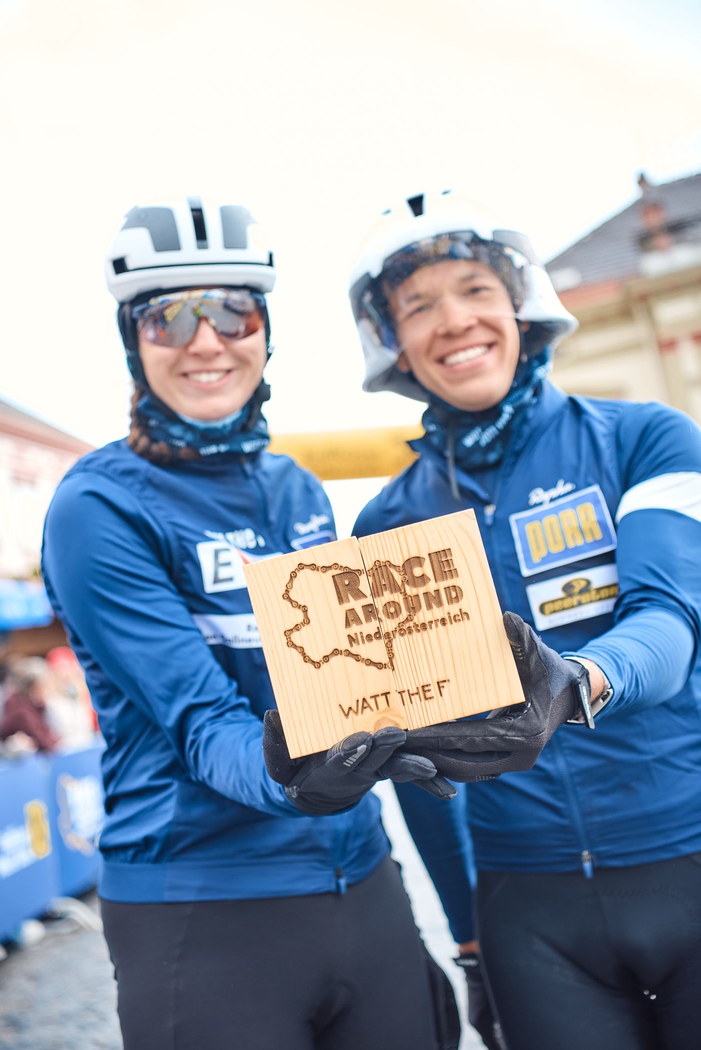Dani und Martin nebeneinander in Teamjacken und Helmen, zwischen ihnen die Holzscheibe mit dem eingebrannten Race-Around-Niederösterreich-Schriftzug